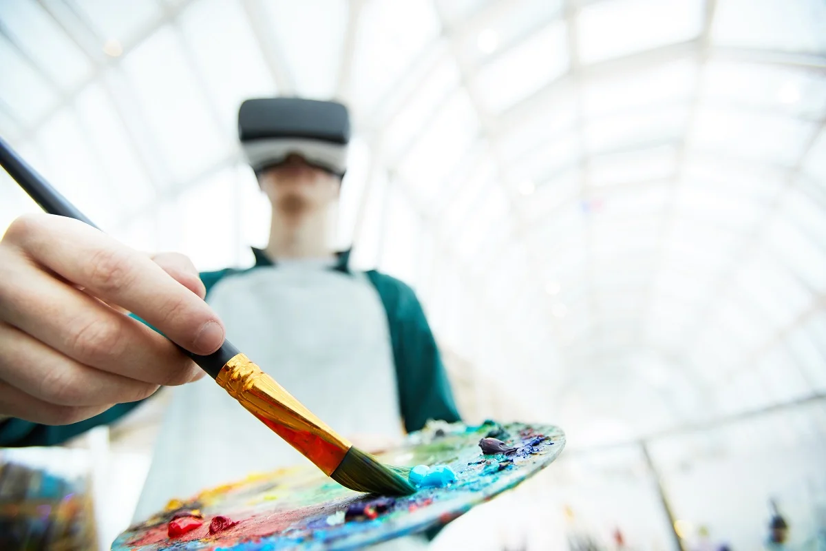 Low angle portrait of young man wearing VR headset painting picture and mixing colors in palette under futuristic glass roof, copy space. VR Painting.