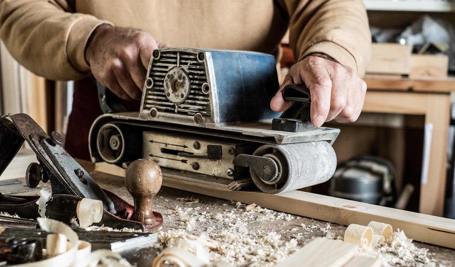 Electric belt sander, sanding machine in male hand. Processing of workpiece on light brown wooden table. Side view, close up