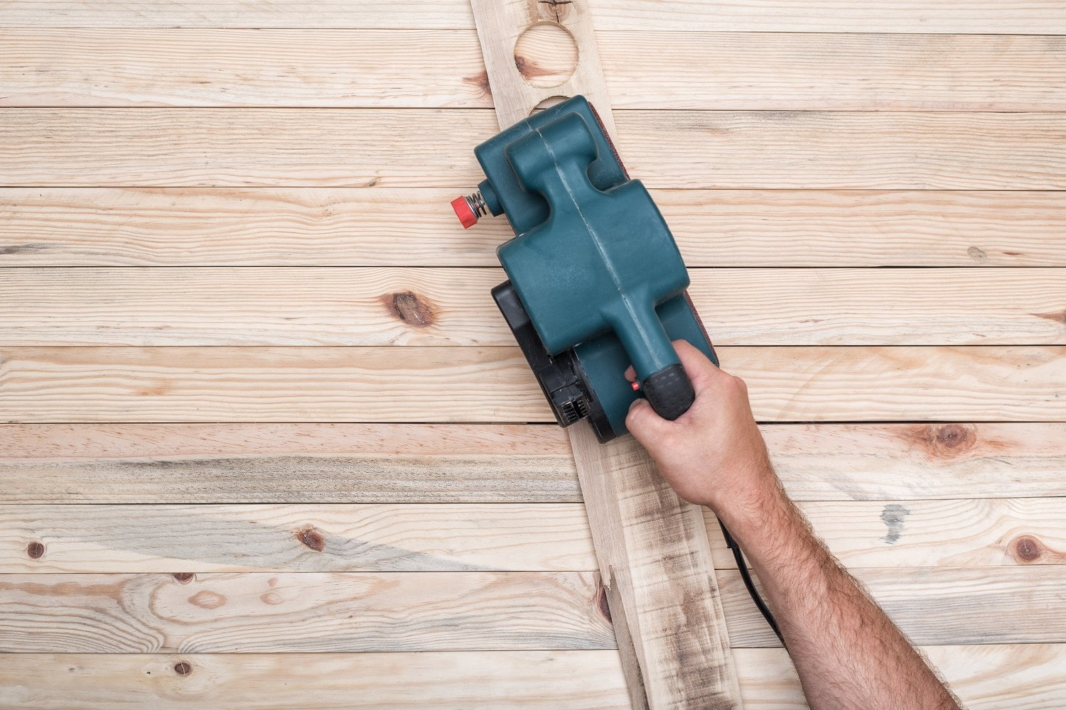 Electric belt sander, sanding machine in male hand. Processing of workpiece on light brown wooden table. Directly above.