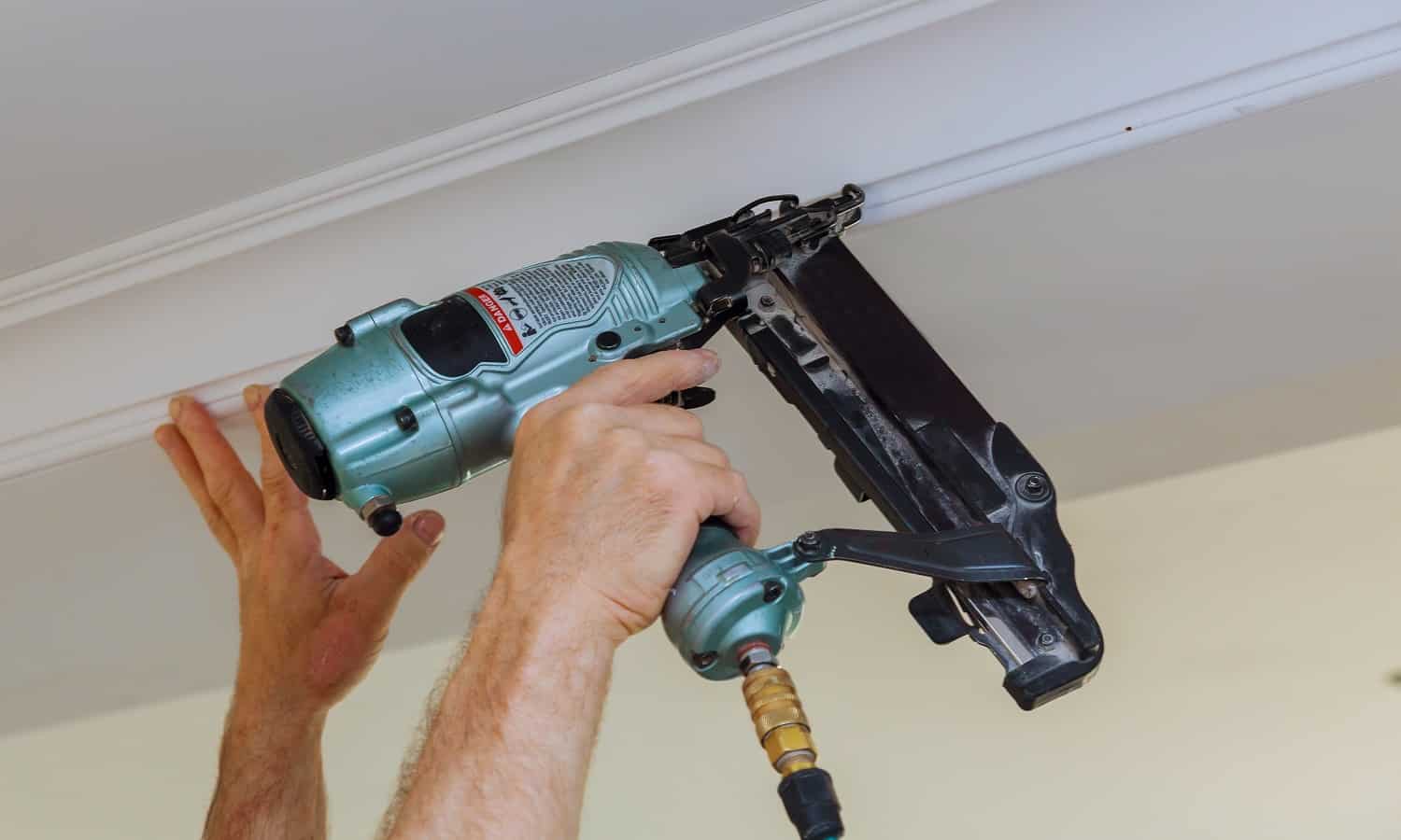 Carpenter brad using nail gun to Crown Moulding framing trim, with the warning label that all power tools have on them shown illustrating safety concept