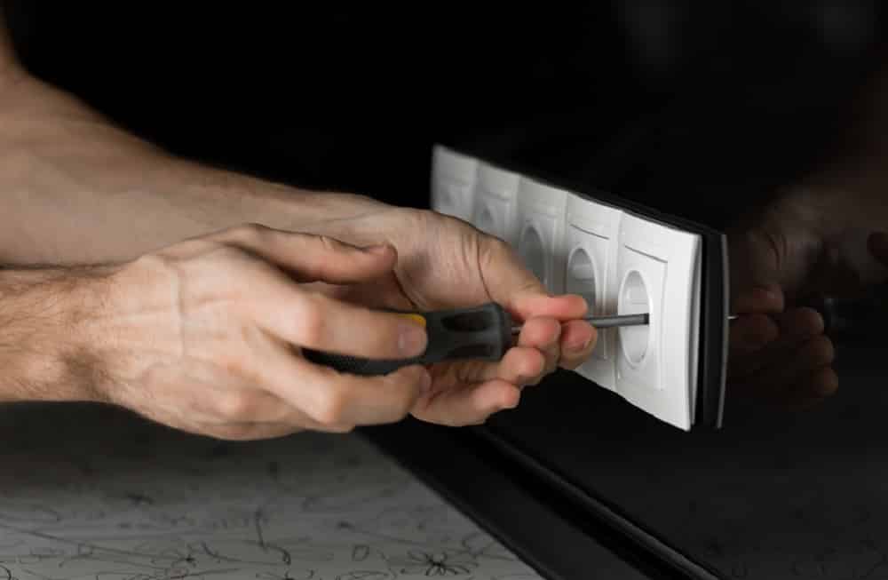 Close-up of an electrician's hand with a screwdriver disassembling a white electrical outlet on a black glass wall.