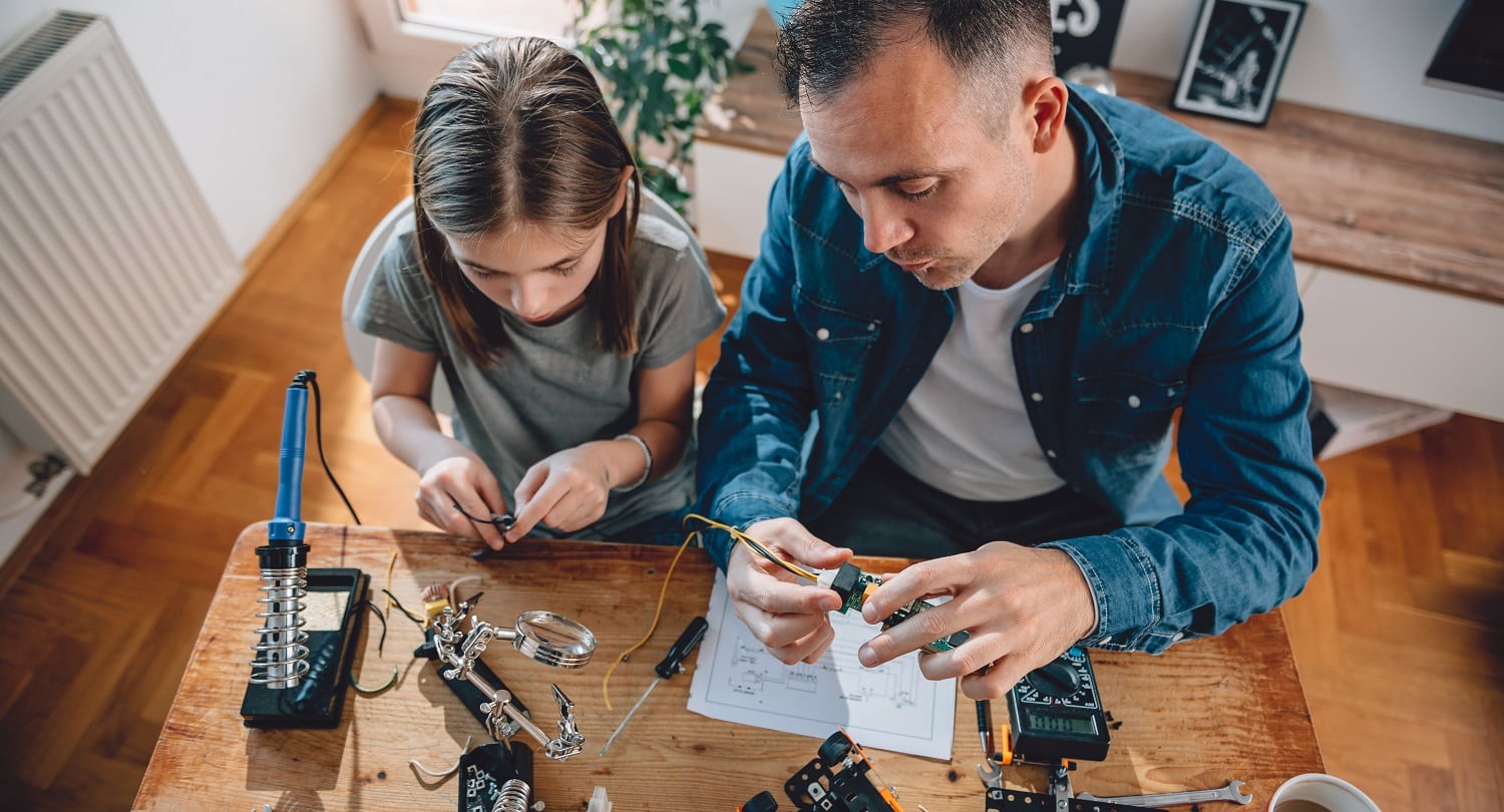 Father and daughter sitting by the wooden table holding circuit board and building robot at home as a school science project