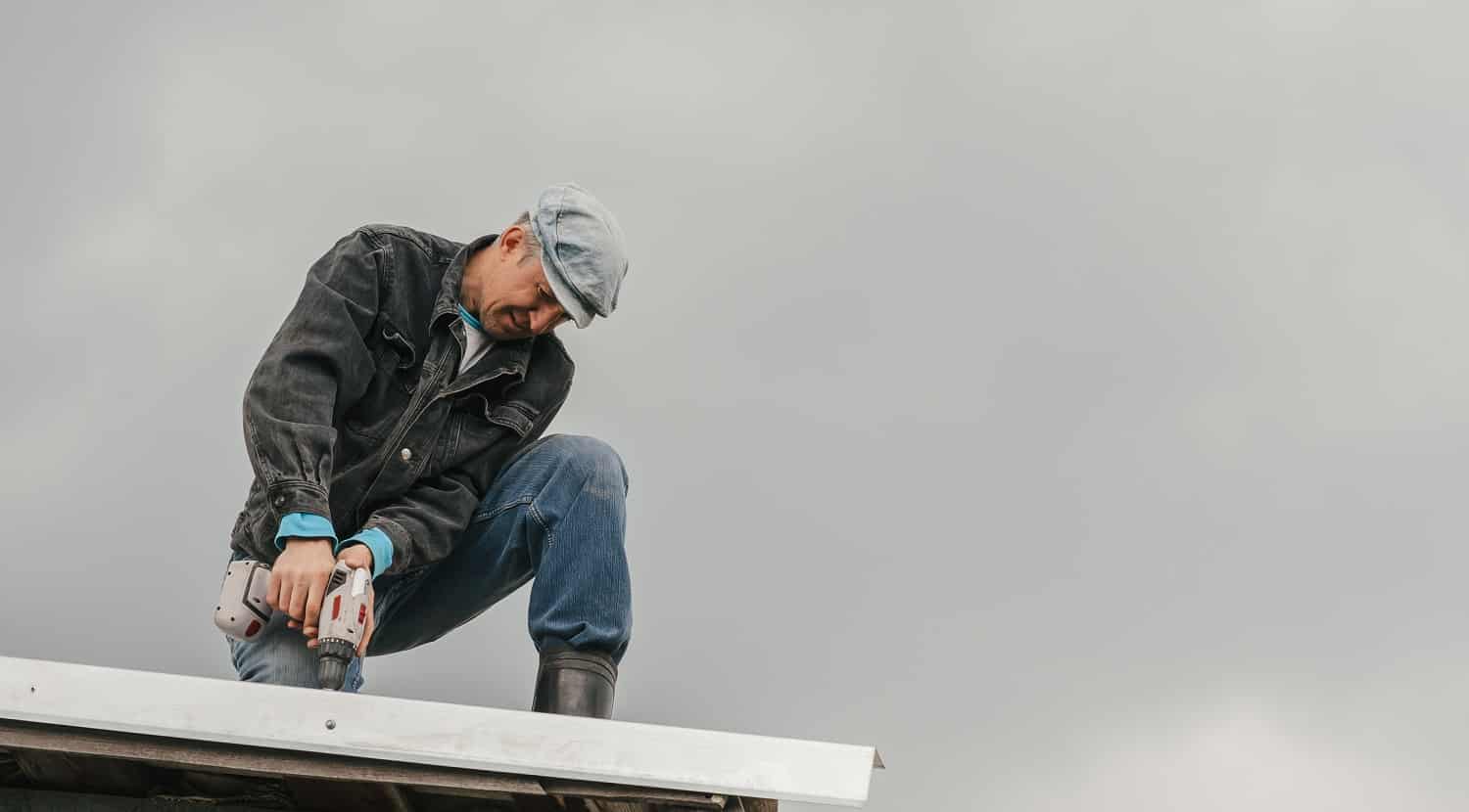 A man in work clothes tighten screws with a screwdriver on the roof against a cloudy sky.