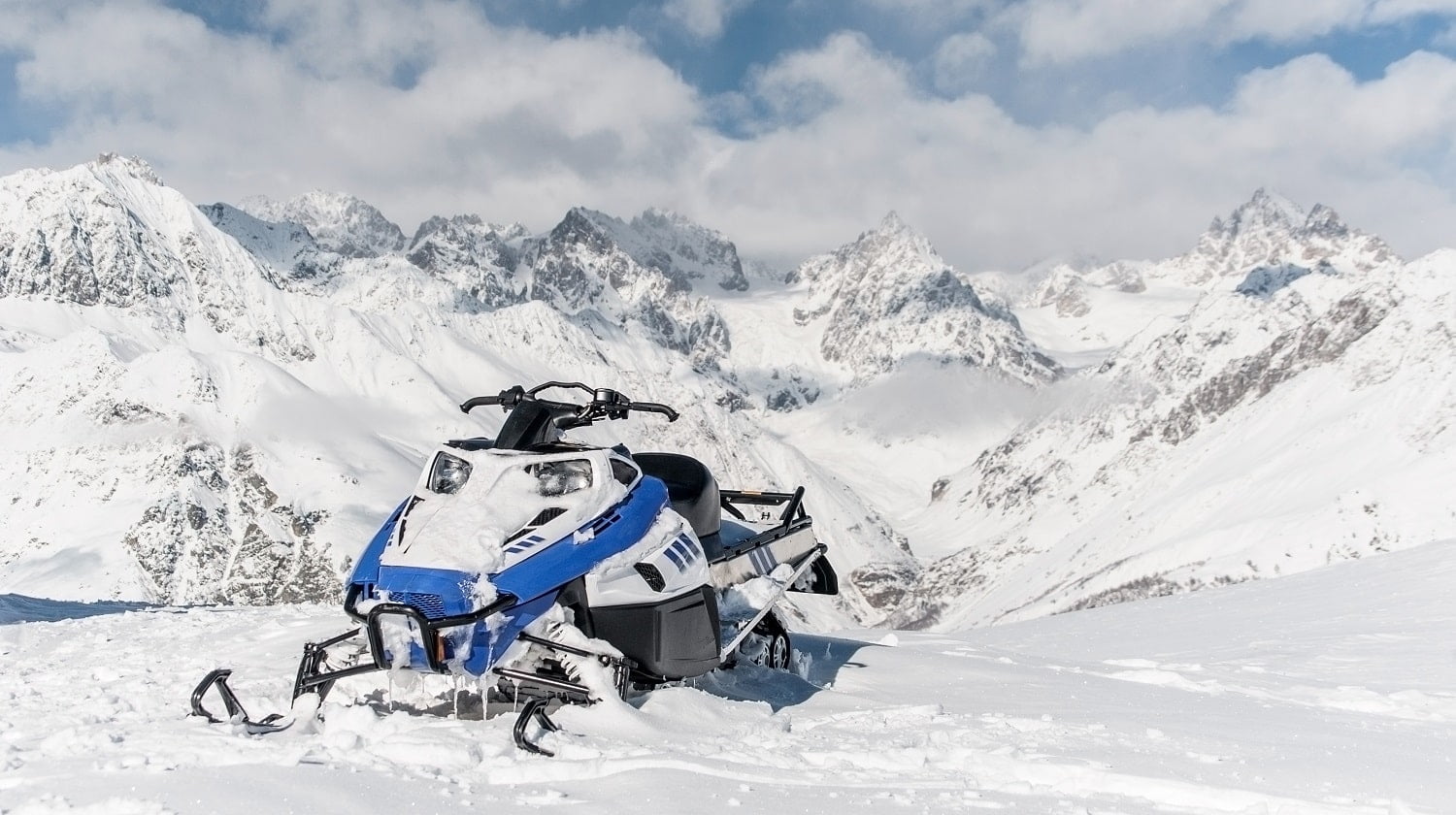 Modern blue snowmobile against the scenic background of high snow mountains peaks and cloudy sky