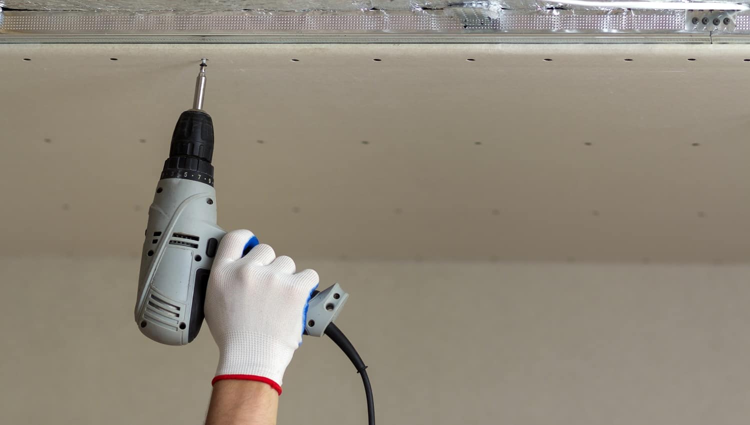 Close-up of constructor worker hand in protective glove with electrical cordless screwdriver connecting suspended drywall ceiling to metal frame. Renovation, construction and DIY concept.