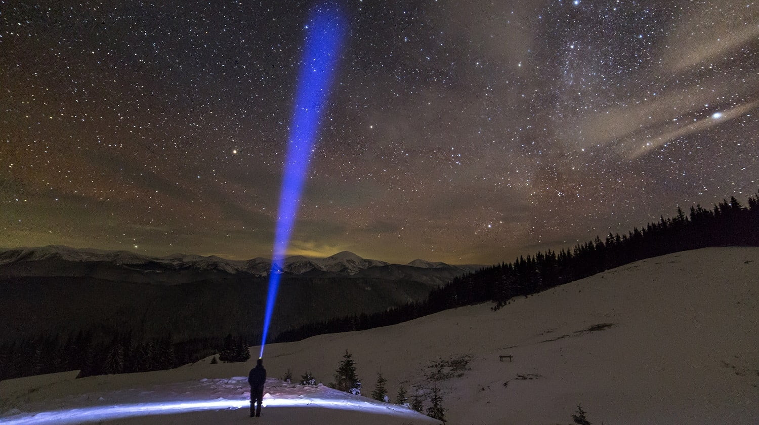 Back view of man with head flashlight standing on snowy valley under beautiful dark blue winter starry sky, bright blue beam on stars and mountains ridge background. Night photography, light painting.