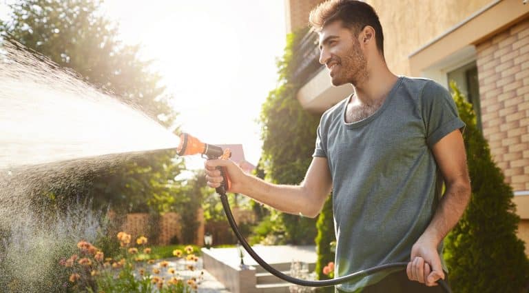 Summer morning in countryside house. Portrait of young attractive tan-skinned bearded man in blue t-shirt smiling, watering plants with hose, working in garden