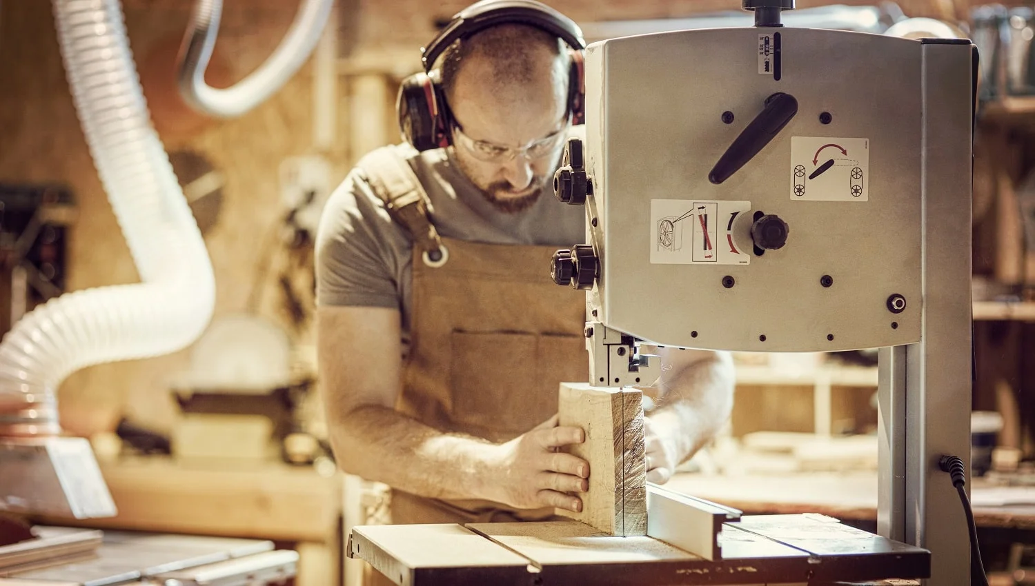 detail of a carpenter at work cutting a plank with a band saw in his workshop, Caucasian with a beard, apron and protective glasses.