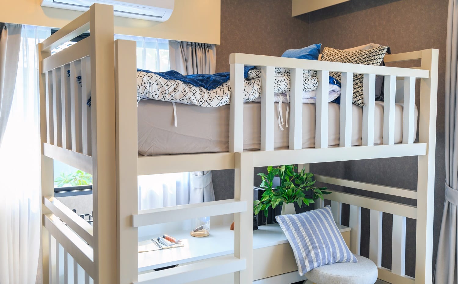 A white wooden bunk bed with a pillow and air conditioner in a children's bedroom with warm light from a window.