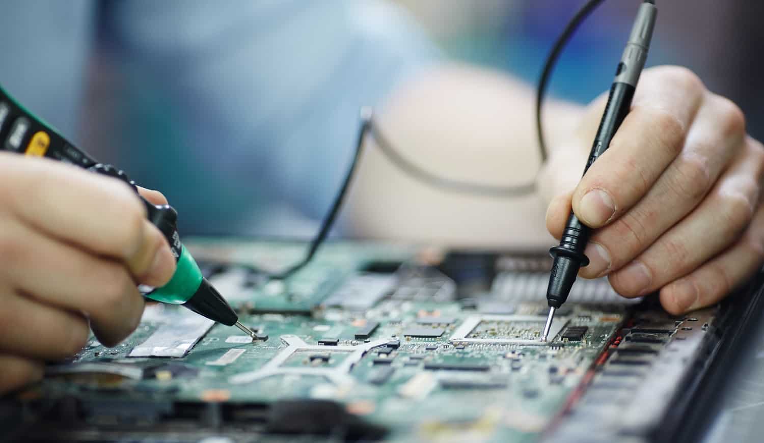 Closeup shot of male hands testing electric current voltage in circuit board of disassembled laptop using multimeter tool on table in maintenance shop
