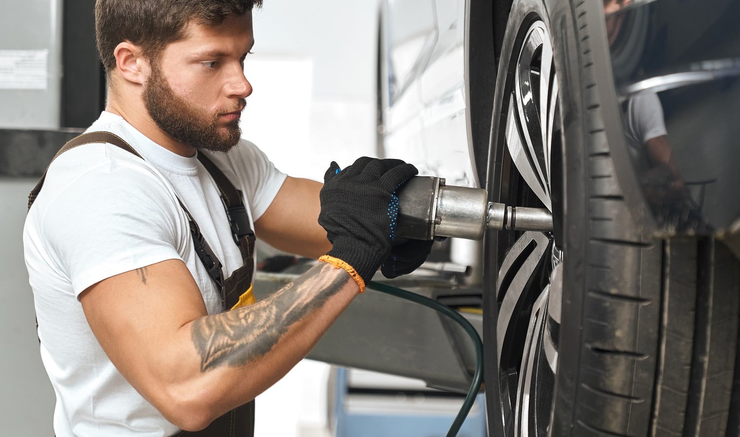 Bearded, brutal mechanic fixing hubcap of auto wheel, using equipment. Muscular man in white t shirt with tattoo on hand wearing in black gloves, working in autoservice.