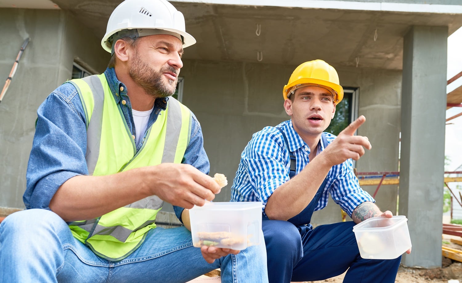 Portrait of two construction workers taking break on site eating lunch out of plastic containers outdoors
