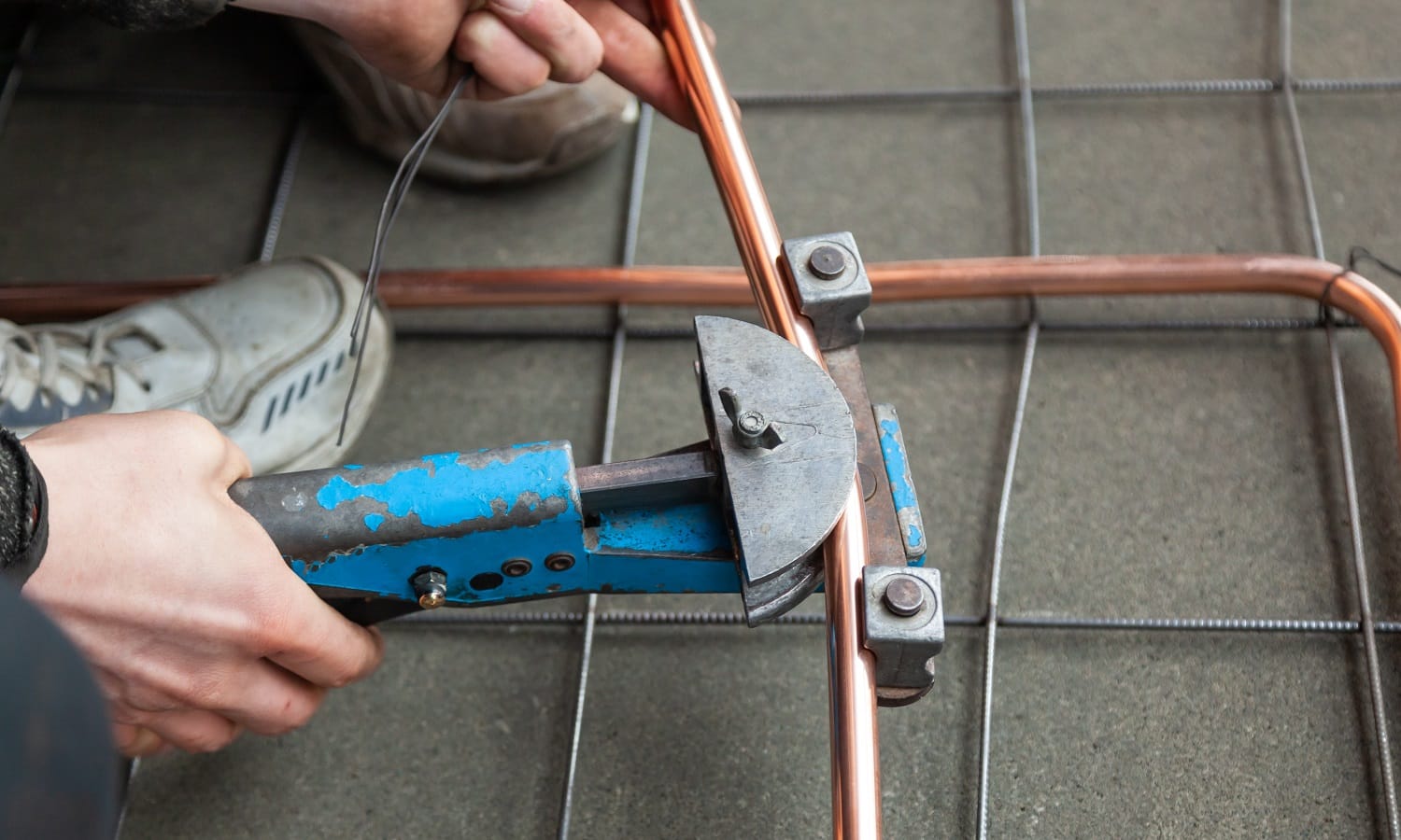 Close-up male hands bends new copper pipes by blue pipe bender, lay pipeline, installation tubes on floor with warm heating of apartment, view from above, horizontal