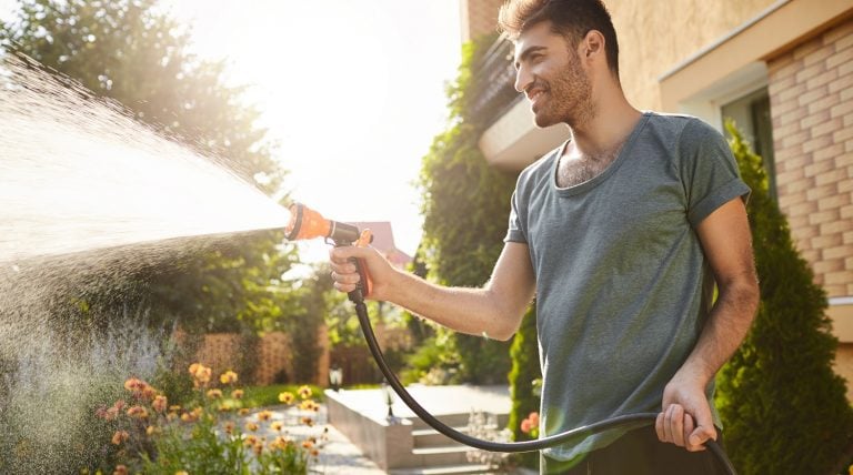 Summer morning in countryside house. Portrait of young attractive tan-skinned bearded man in blue t-shirt smiling, watering plants with hose, working in garden