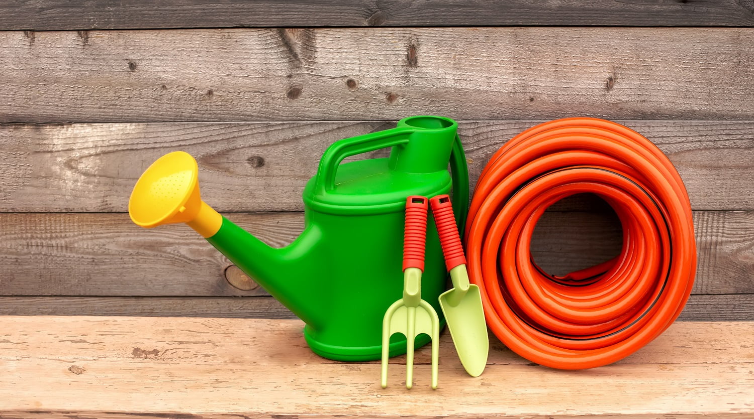 Green garden watering can, orange watering hose and small green hand gardening tools stand on wooden background. Copy space