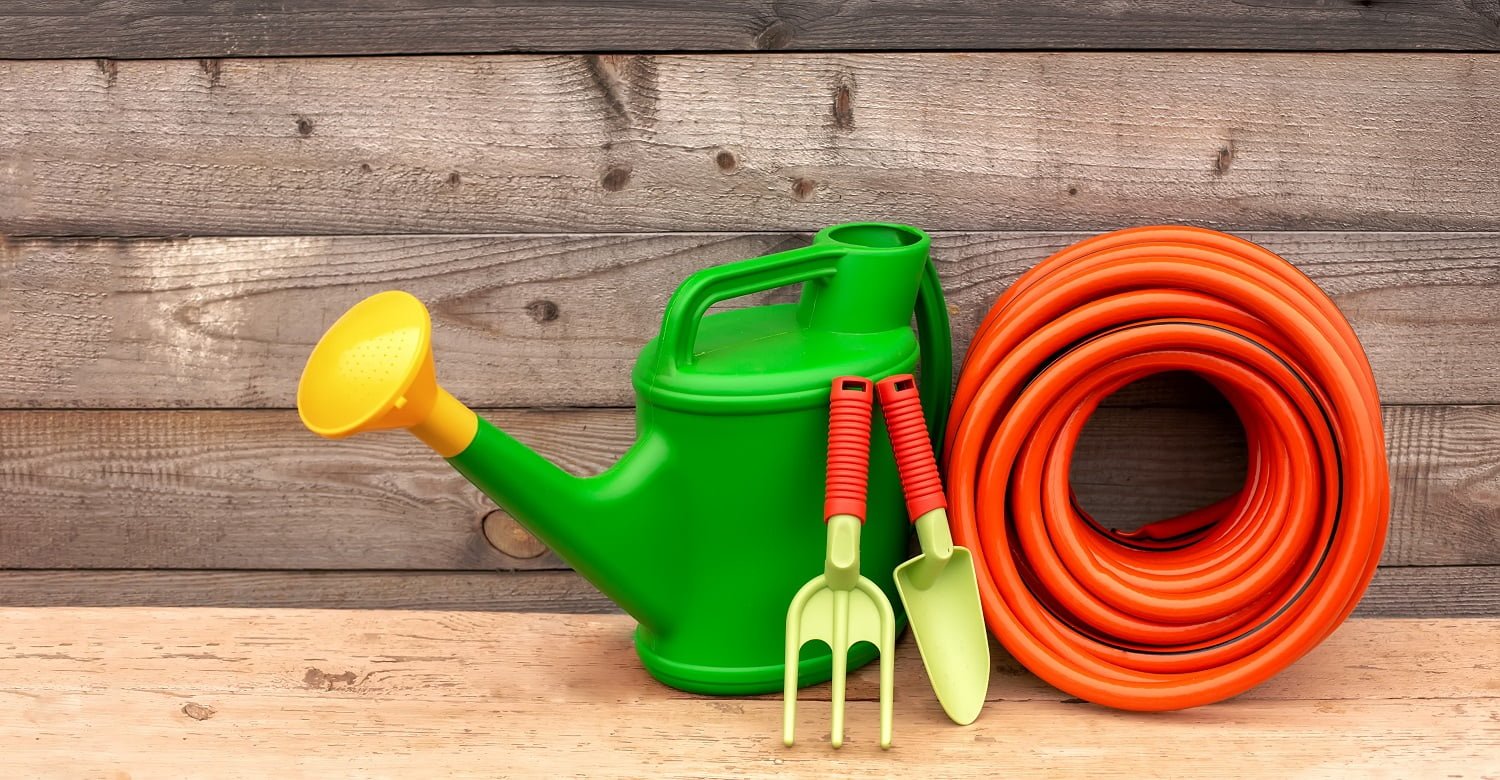 Green garden watering can, orange watering hose and small green hand gardening tools stand on wooden background. Copy space