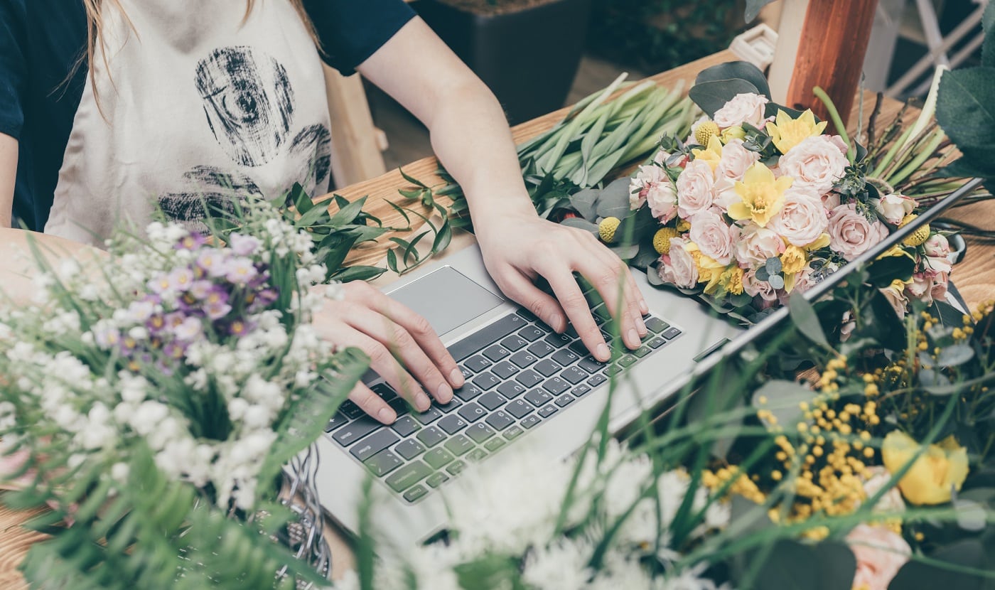 Faceless view of saleswoman in apron browsing laptop sitting at table with flowers in market.