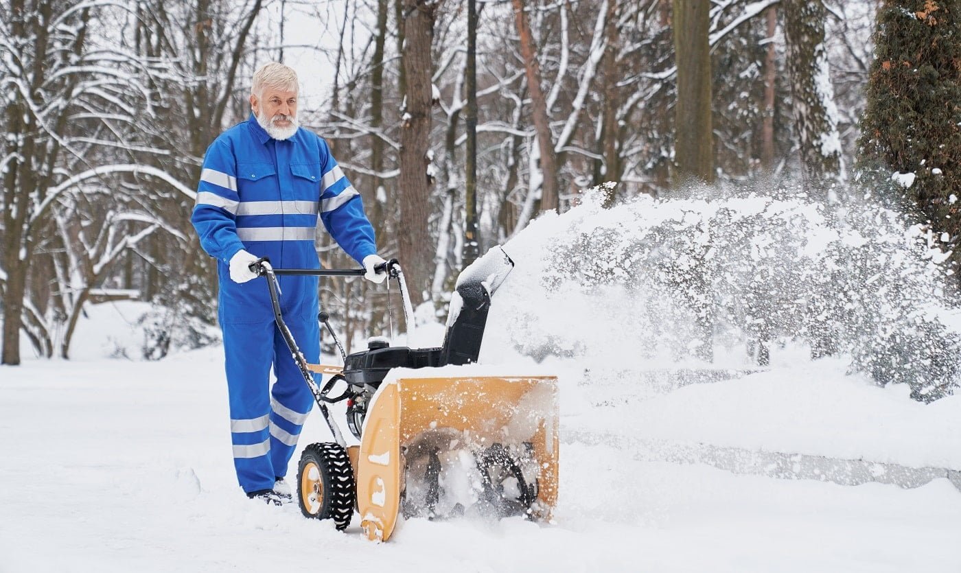 View from side bearded man wearing blue uniform working with snowblower and removing snow from footpath. Elder man keeping machine and driving in park. Concept of city service and maintenance.