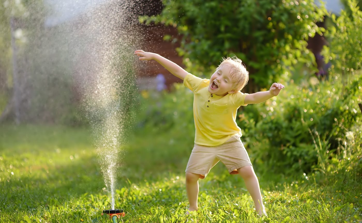 Funny little boy playing with garden sprinkler in sunny backyard. Preschooler child having fun with spray of water. Summer outdoors activity for kids.