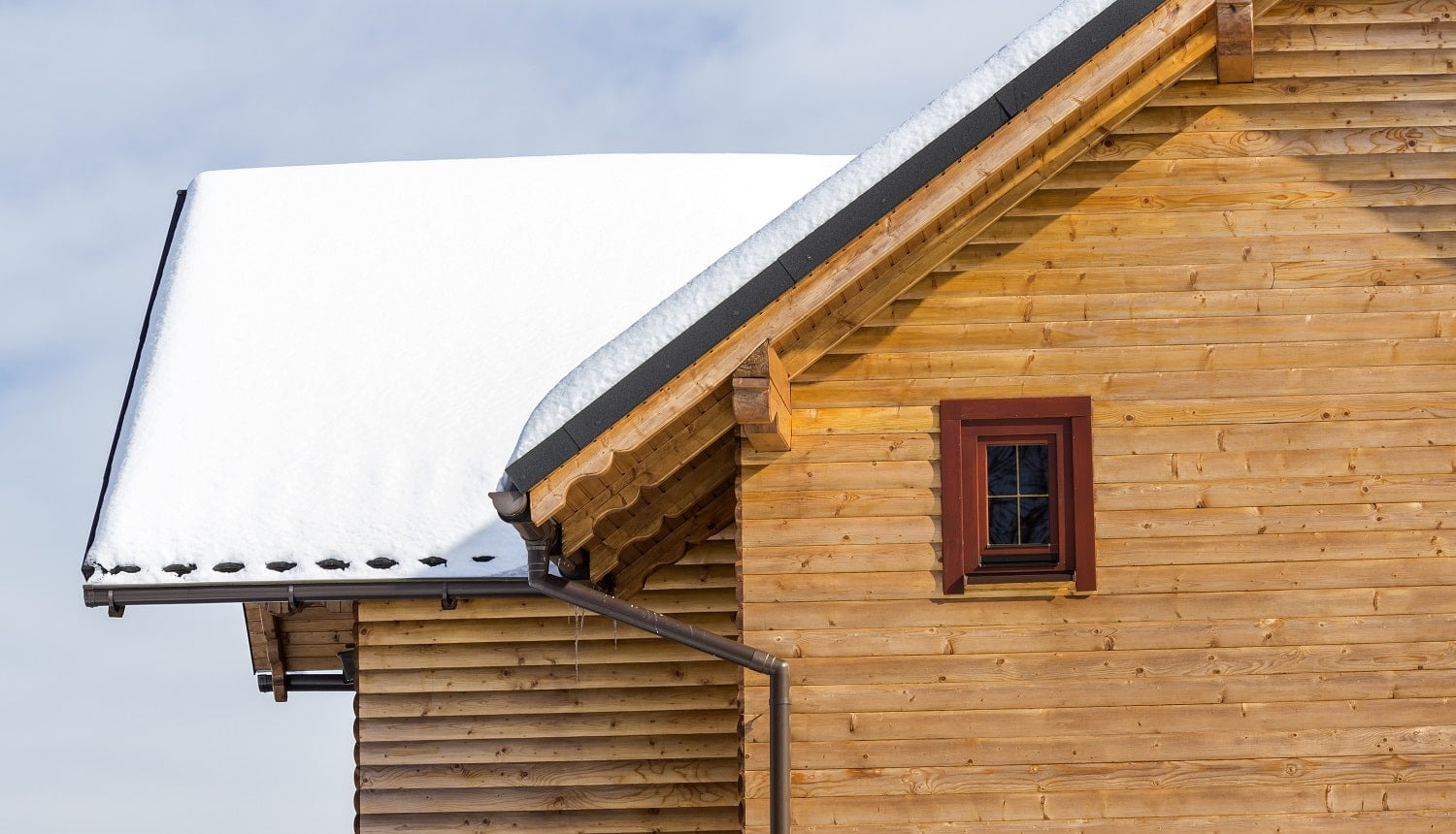 Detail top of wooden ecological traditional cottage of lumber materials with steep roof, attic rooms covered with snow on sunny winter day. Old traditions and modern professional construction concept. EasyOn Gutter Guard Review