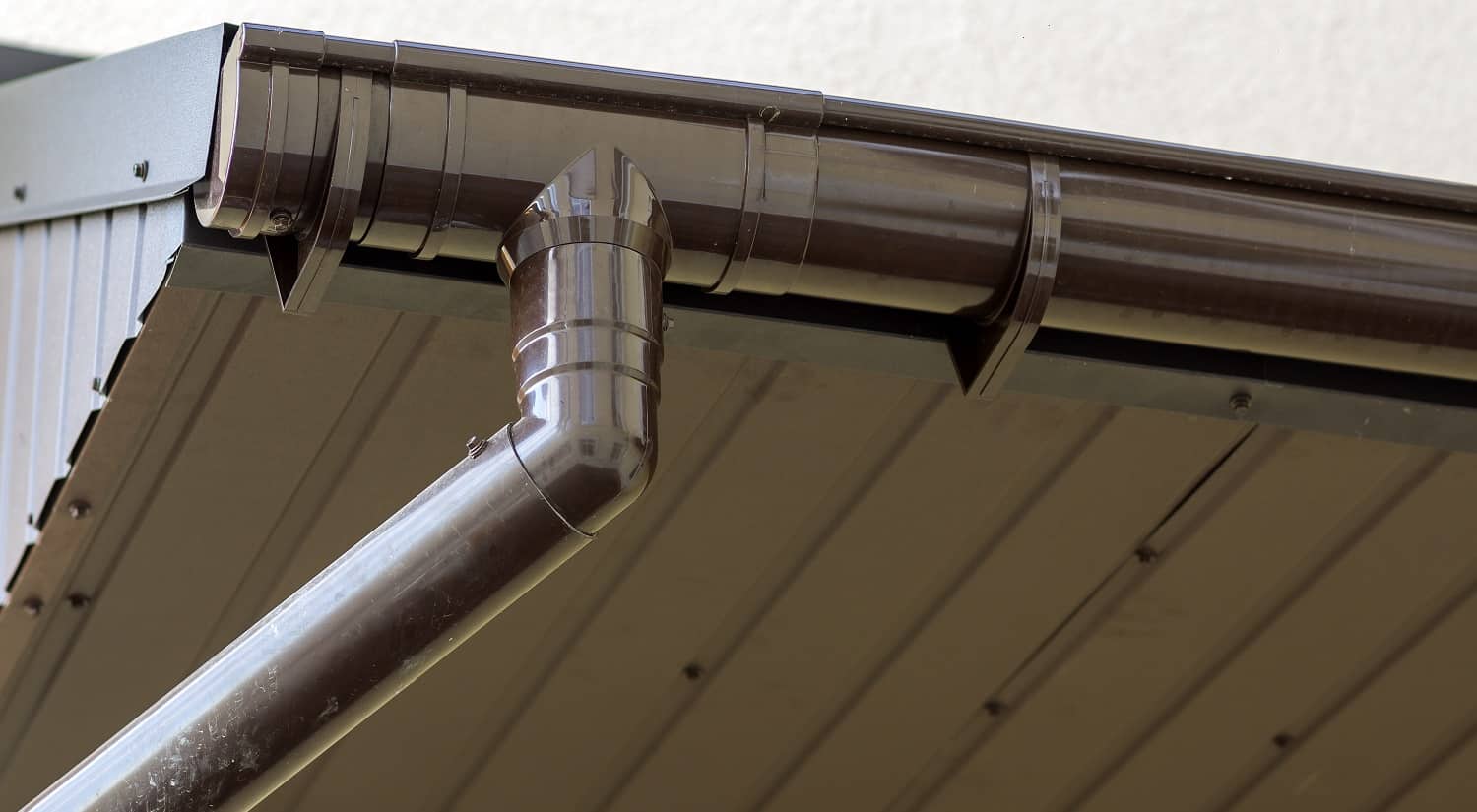 Close-up detail of cottage house corner with brown metal planks siding and roof with steel gutter rain system. Roofing, construction, drainage pipes installation and connection concept.