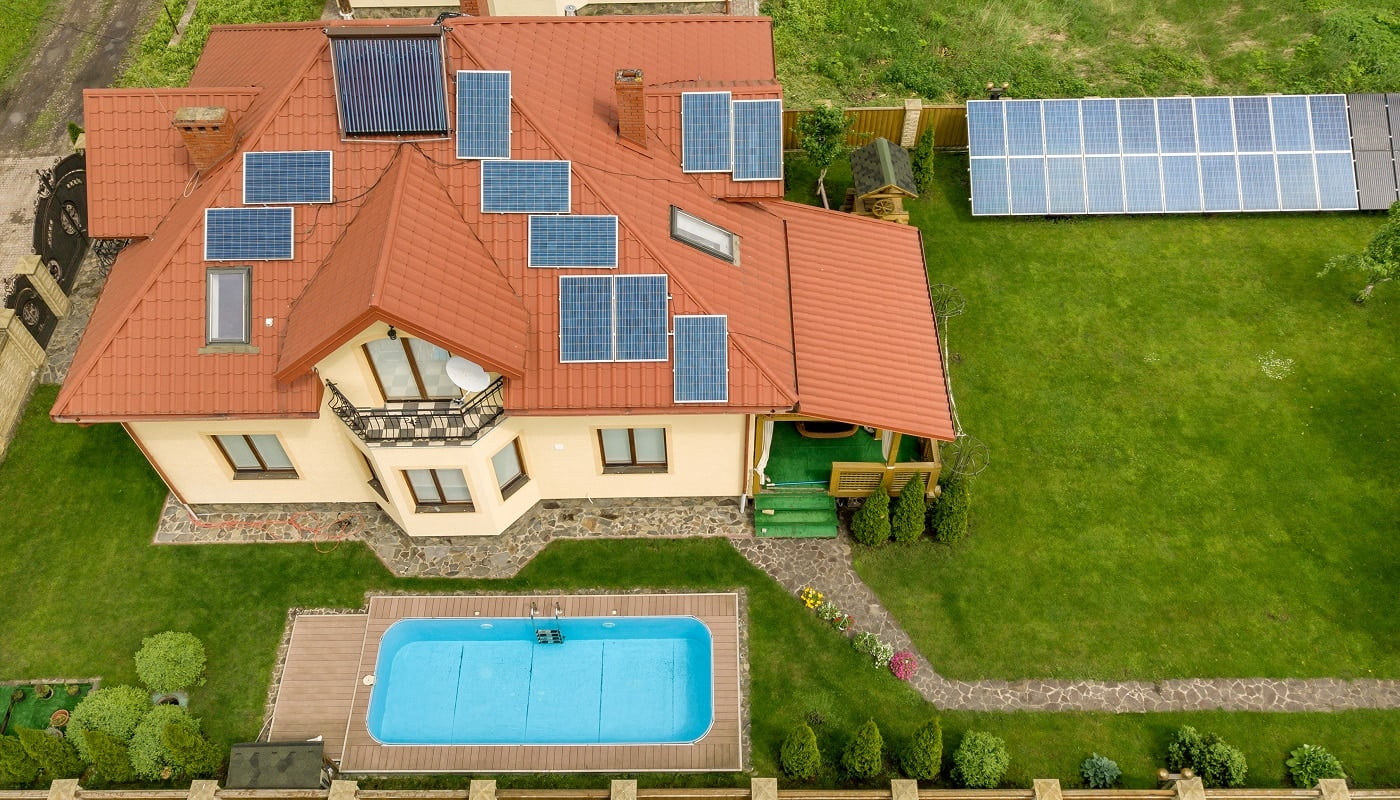 Aerial view of a new autonomous house with solar panels and water heating radiators on the roof and green yard with blue swimming pool.