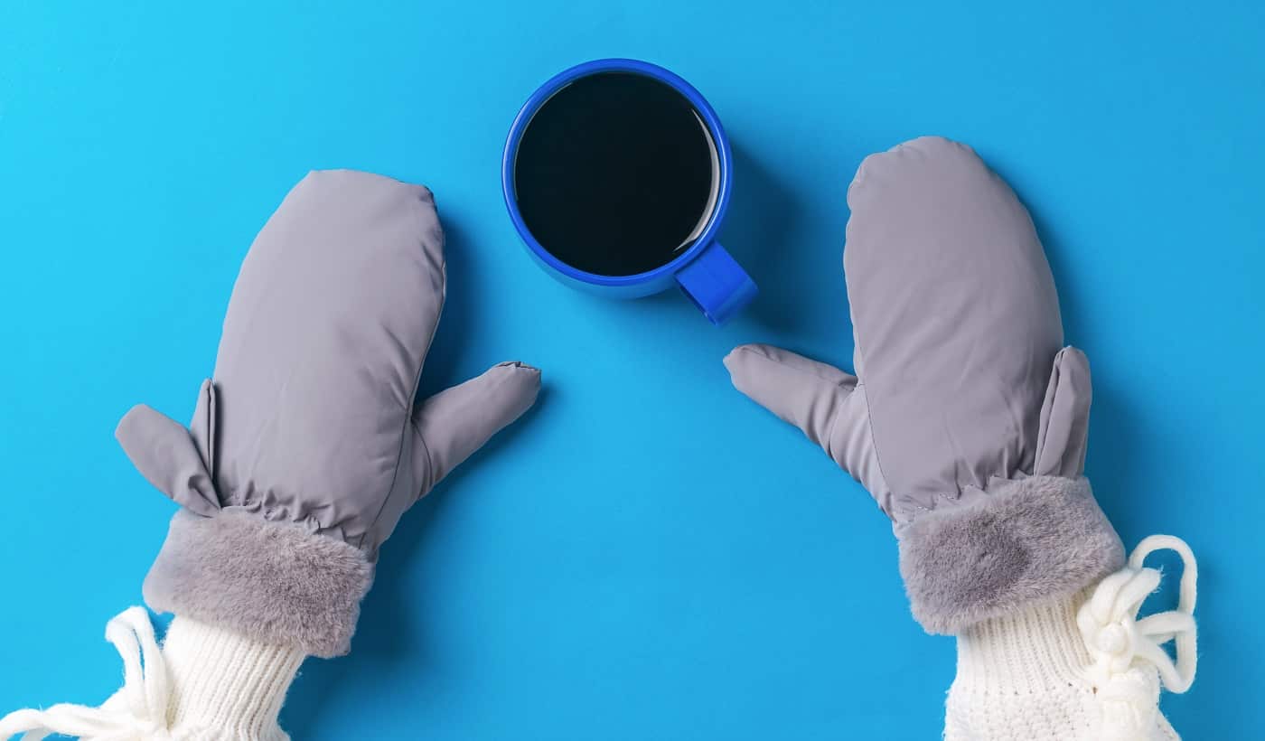 A girl's hands in mittens and a cup of black coffee on a blue background. Hot drink and Best Warmest Mittens