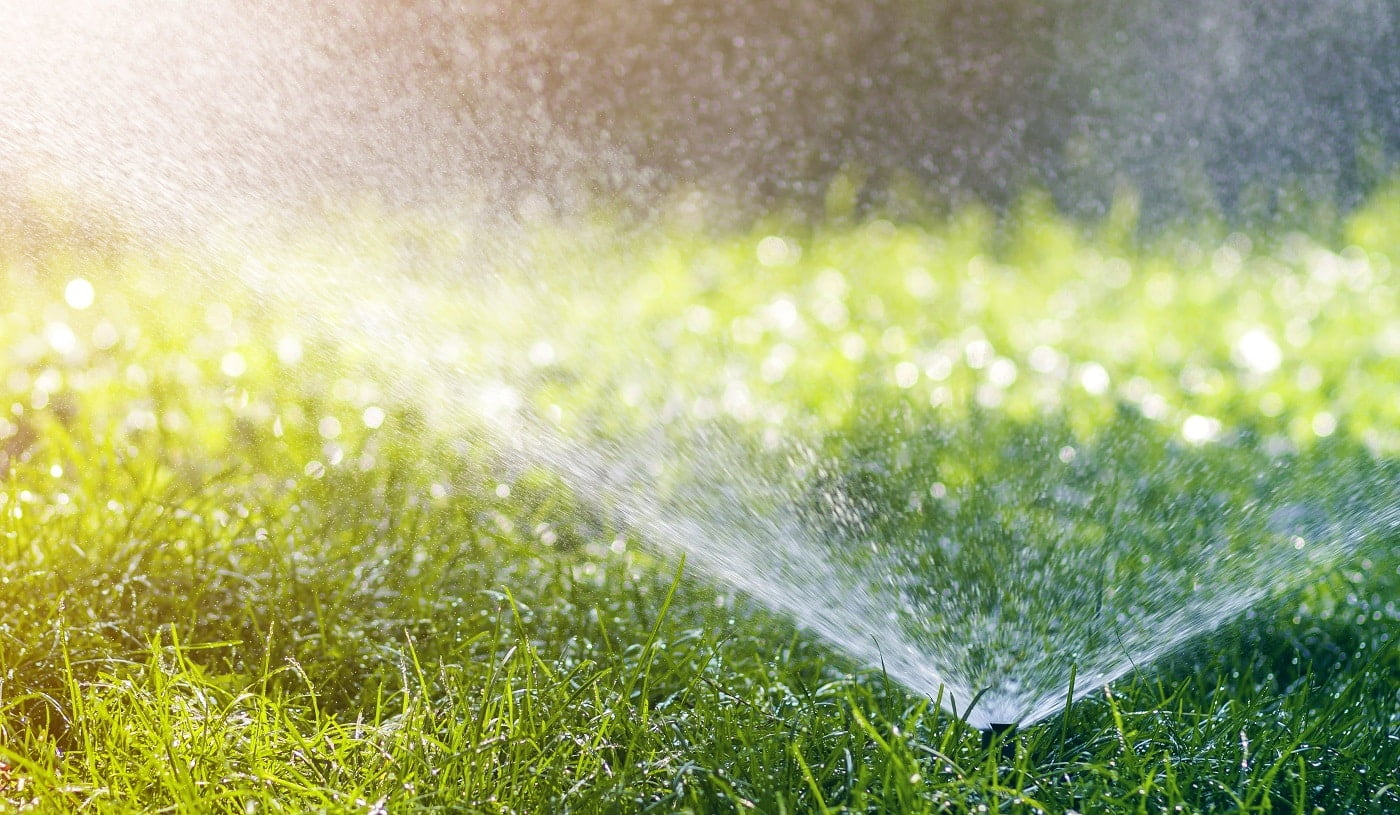 Lawn water sprinkler spraying water over lawn green fresh grass in garden or backyard on hot summer day. Automatic watering equipment, lawn maintenance, gardening and tools concept.