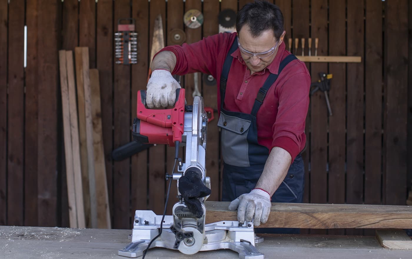 A man in protective overalls and glasses cuts a wooden beam using a circular saw. Home furniture workshop. Artisan works in his carpentry workshop.