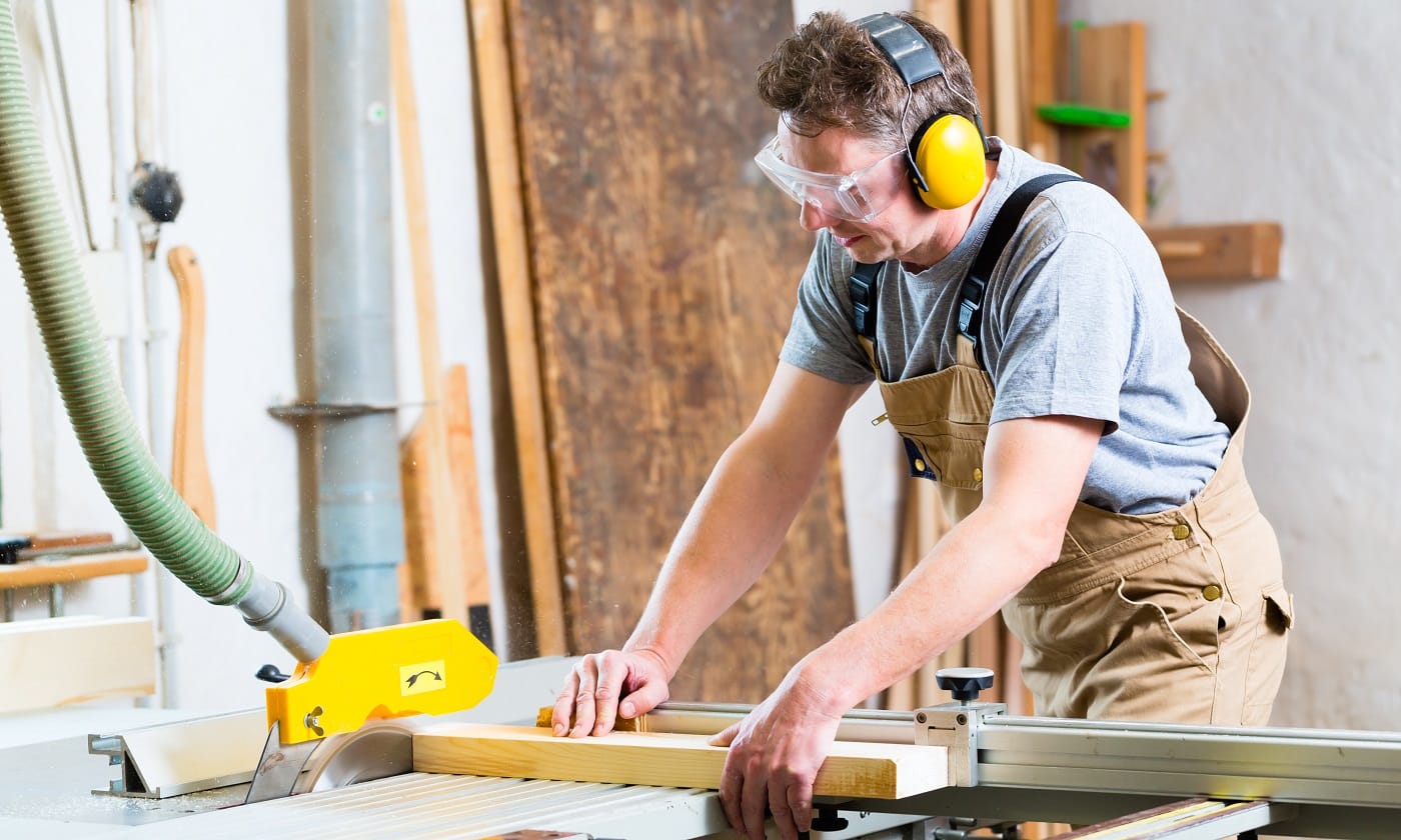 Carpenter working on an electric buzz saw cutting some boards, he is wearing safety glasses and hearing protection for workplace safety