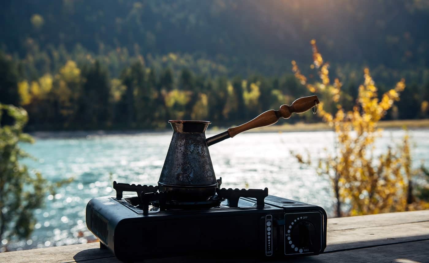 Copper turk with coffee on gas burner on the background of river and hills. Coffee preparing on the banks of mountain river on portable gas stove during hiking trip. Tourism concept.