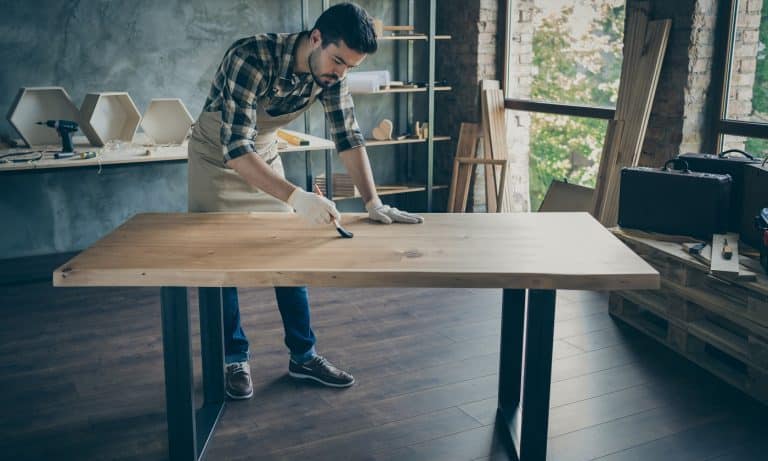 Full length photo of handsome careful guy covering handmade slab table oil varnish paint brush own wooden business industry woodwork shop garage indoors