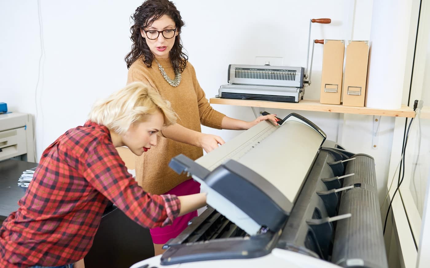 Portrait of two women setting up plotter machine in printing shop, focus on manager giving instructions to employee, copy space