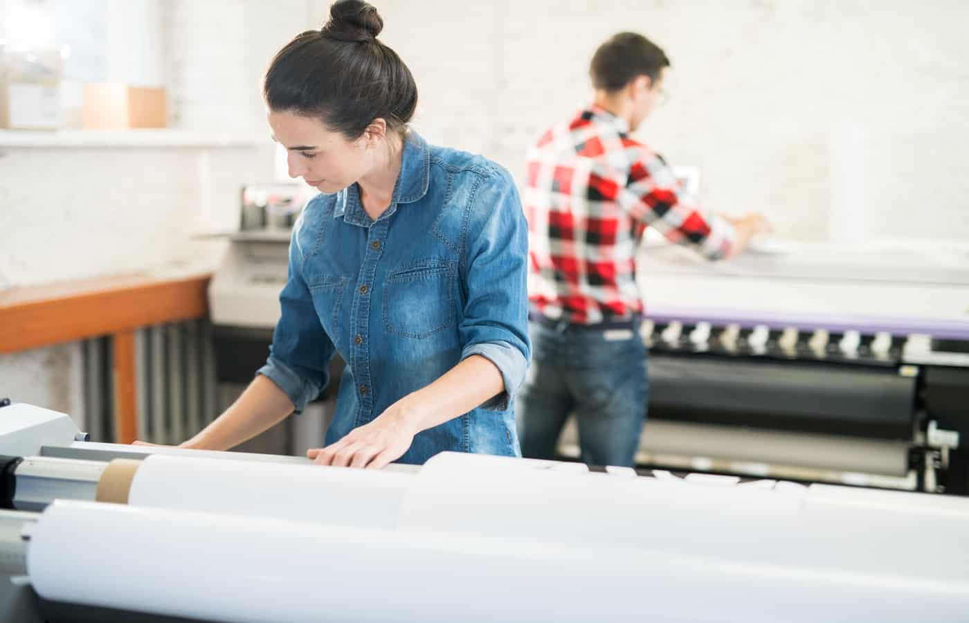 Serious concentrated woman in denim shirt standing at large format printer and choosing program while working in printing office