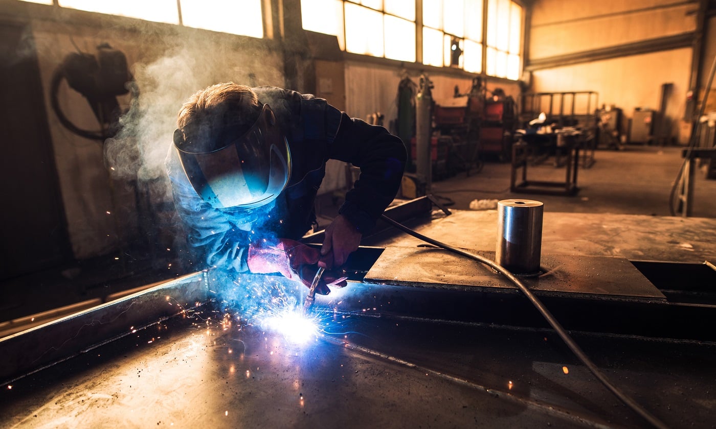 Professional welder in protective uniform and helmet welding metal part in workshop.