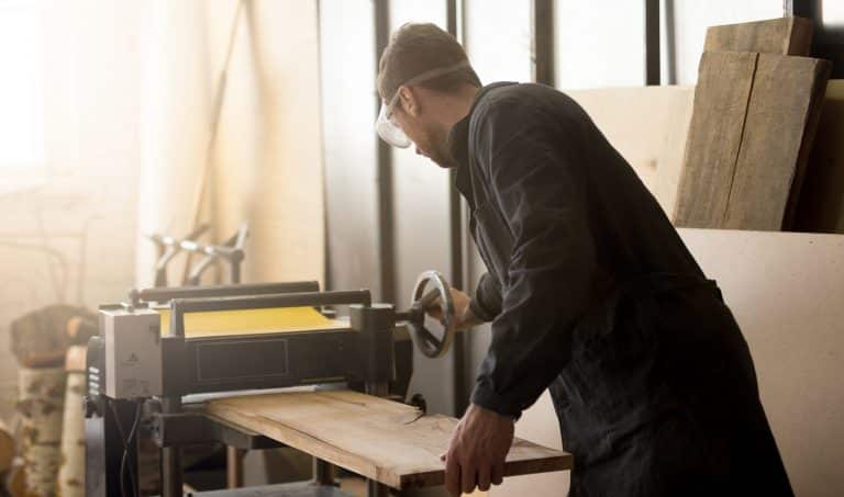 Back view of young carpenter or craftsman wearing working clothes and protective eyewear woodworking in small workshop interior, operating stationary power planer, processing trimming wooden plank DeWalt DW735X Planer