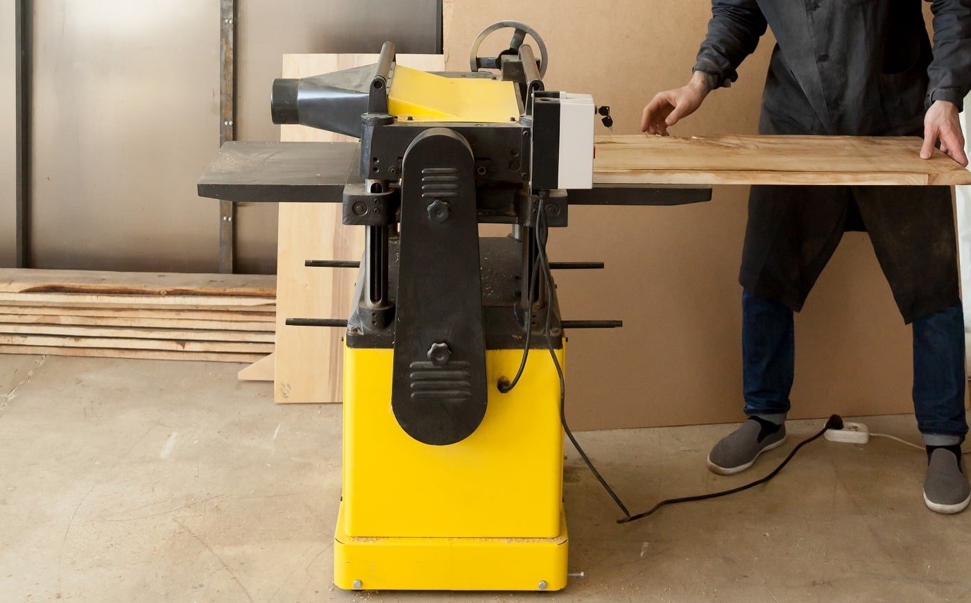 Worker in safety glasses putting untreated wooden board in thickness planer machine. Carpenter producing lumber with powerful machine tool in sawmill. Skilled joiner working in woodworking workshop