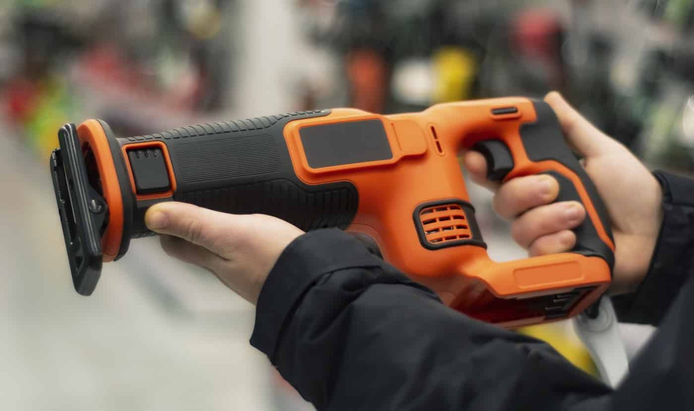 Man holds an orange reciprocating saw for repair work against the backdrop of showcases in a hardware store