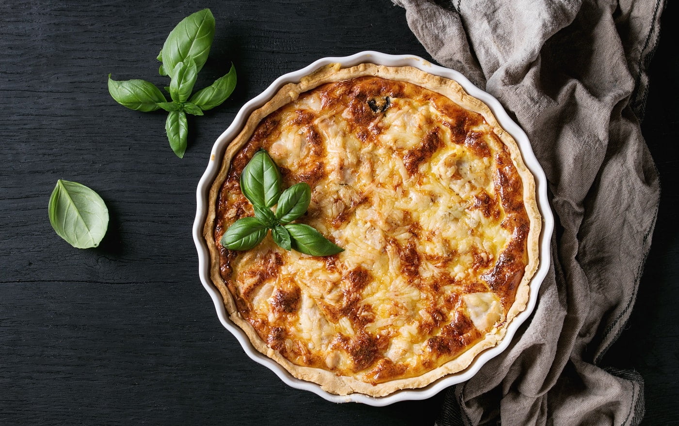 Baked homemade quiche pie in white ceramic form served with fresh greens and textile towel on black wooden burned background. Flat lay with copy space