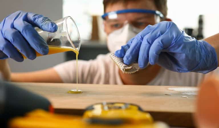 Close-up of carpenter hands in blue gloves working at workshop. Man in respirator and glasses pouring wood varnish on wooden surface. Carpentry work and joinery concept