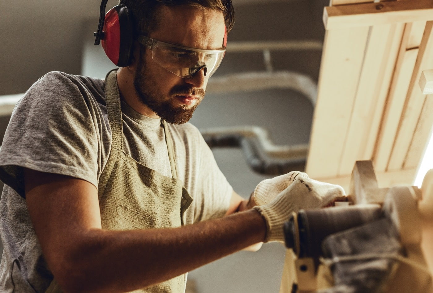Handsome bearded guy in protection goggles and earmuffs making small timber part on belt sander in workshop
