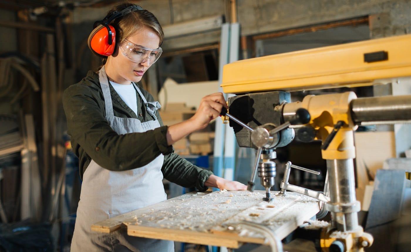 Pretty young carpenter wearing ear and eye protectors while using drill press to make holes in wooden work piece, waist-up portrait