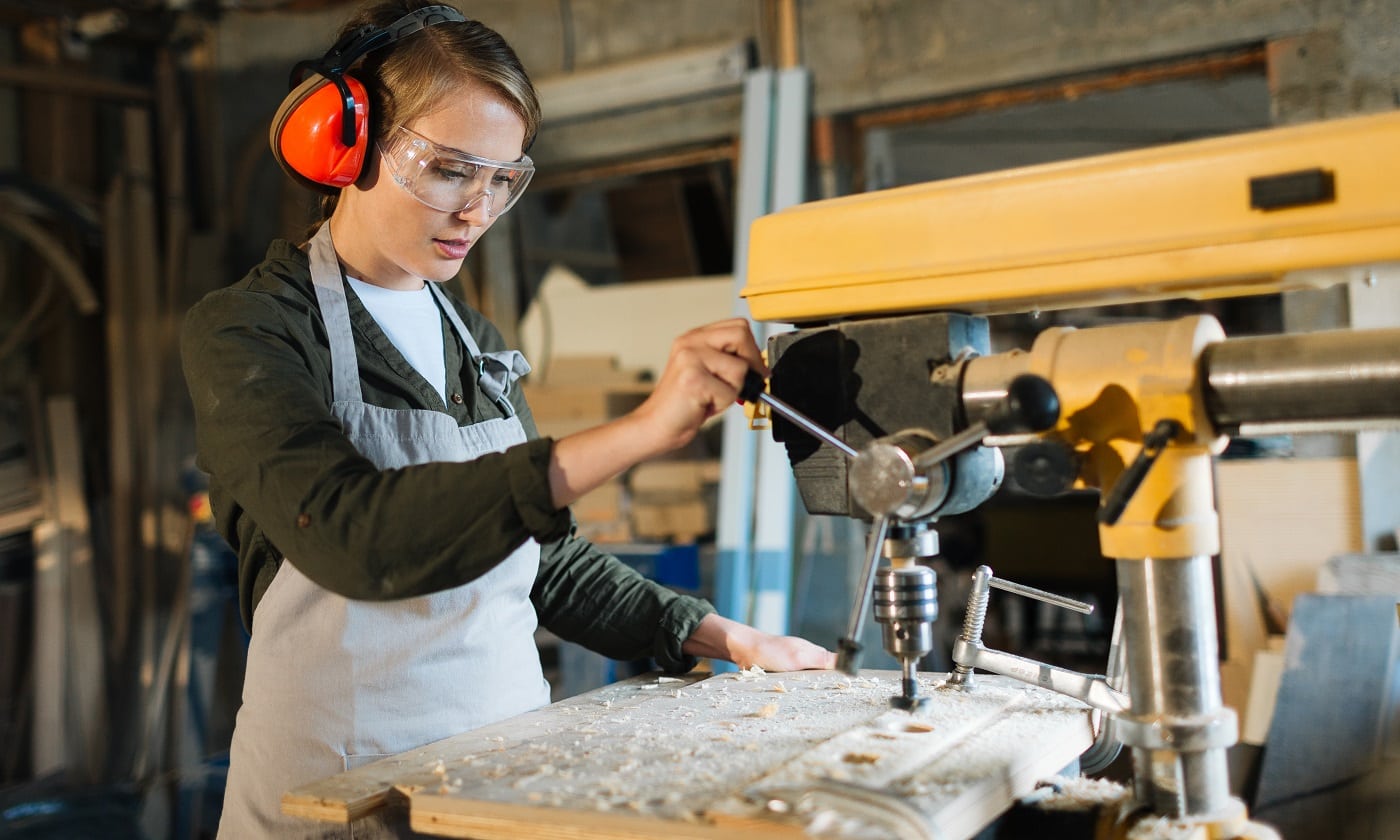 Pretty young carpenter wearing ear and eye protectors while using drill press to make holes in wooden work piece, waist-up portrait