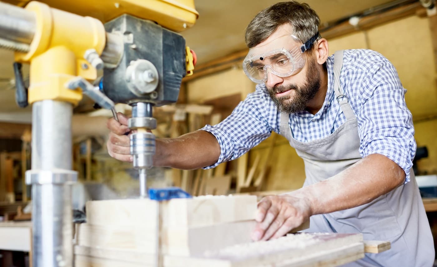 Handsome bearded carpenter wearing safety goggles and apron using drill press machine in order to make holes in wooden plank, interior of spacious workshop on background