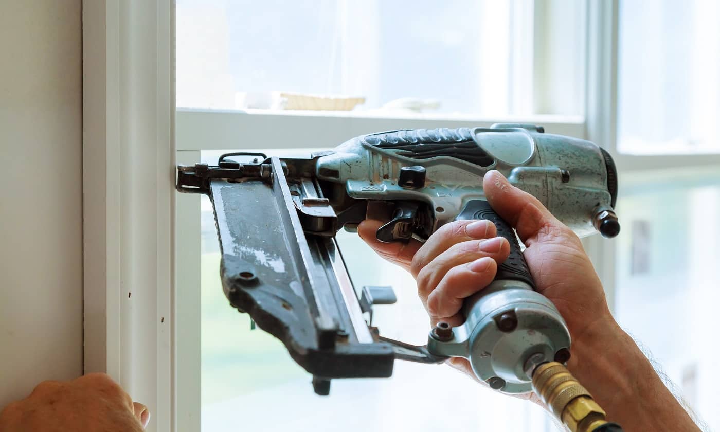 Carpenter brad using nail gun to moldings on windows, framing trim, with the warning label that all power tools have on them shown illustrating safety concept. Framing Nailer Verdict