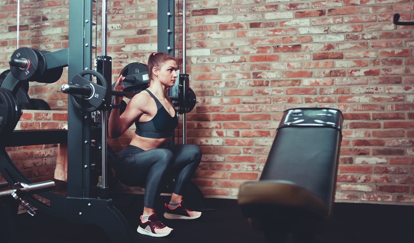 Concentrated sporty girl exercising squatting with barbell at smith machine against brick wall in gym