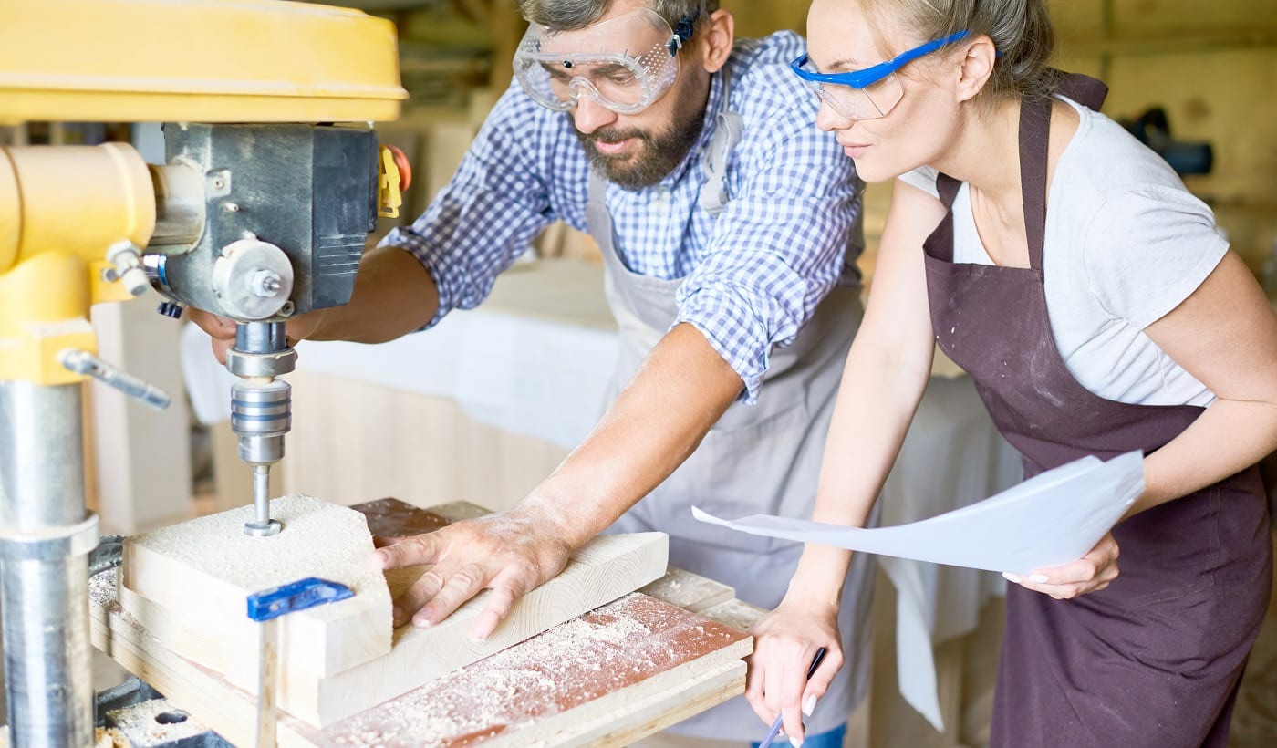 Concentrated bearded carpenter explaining his attractive apprentice how to use drill press machine, interior of spacious workshop on background