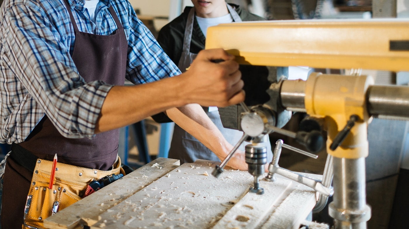 Skillful woodworker in eye and ear protectors explaining his female apprentice how to use drill press, she listening to him with attention