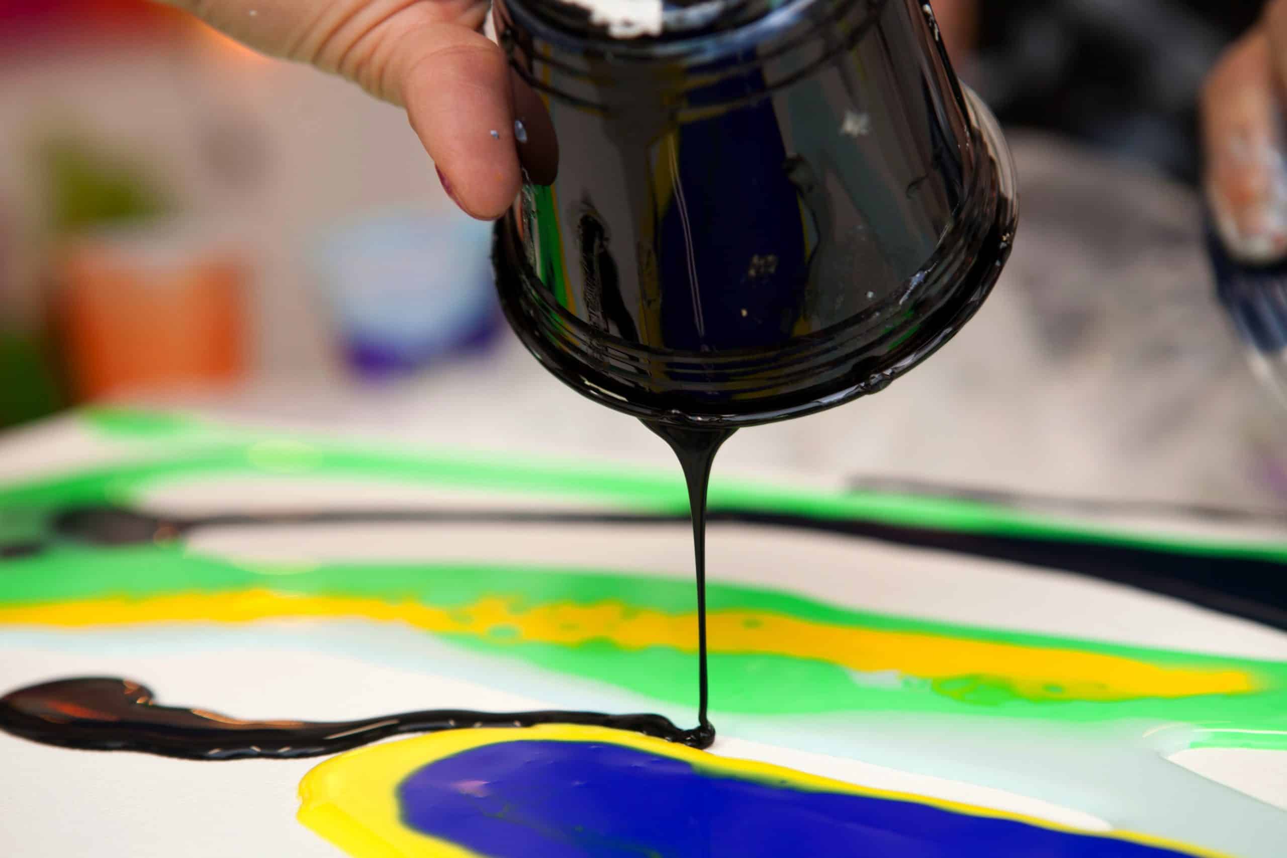 A person pours a small stream of black paint from a plastic cup onto a canvas with swirls of green neon and yellow during the process of fluid painting with acrylic paints mixed with floetrol medium.; Shutterstock ID 1420128320; Purchase Order: AD126