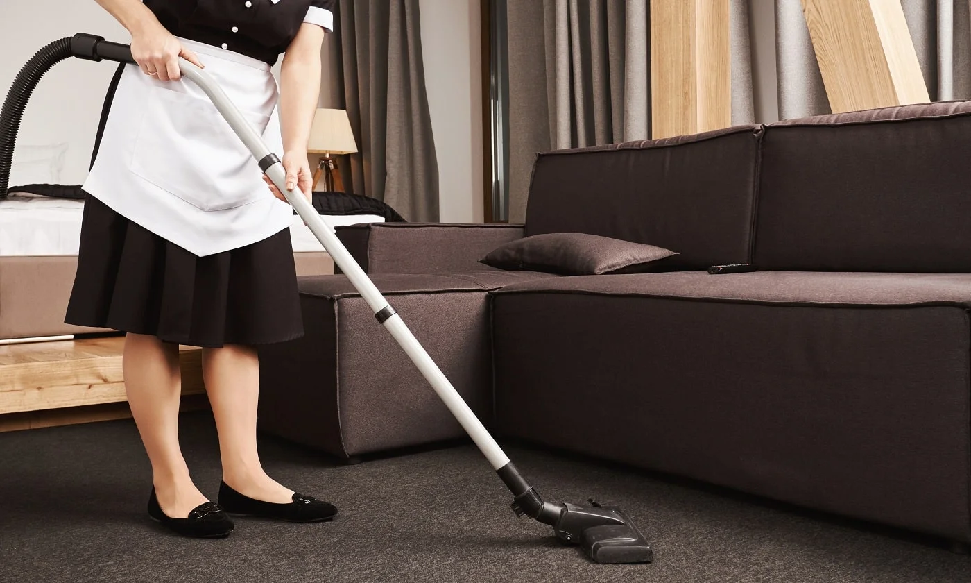 Clean house is key for productivity. Cropped shot of housemaid during work, cleaning living room with vacuum cleaner, removing dirt and mess near sofa. Maid is ready to make this place shine bright.